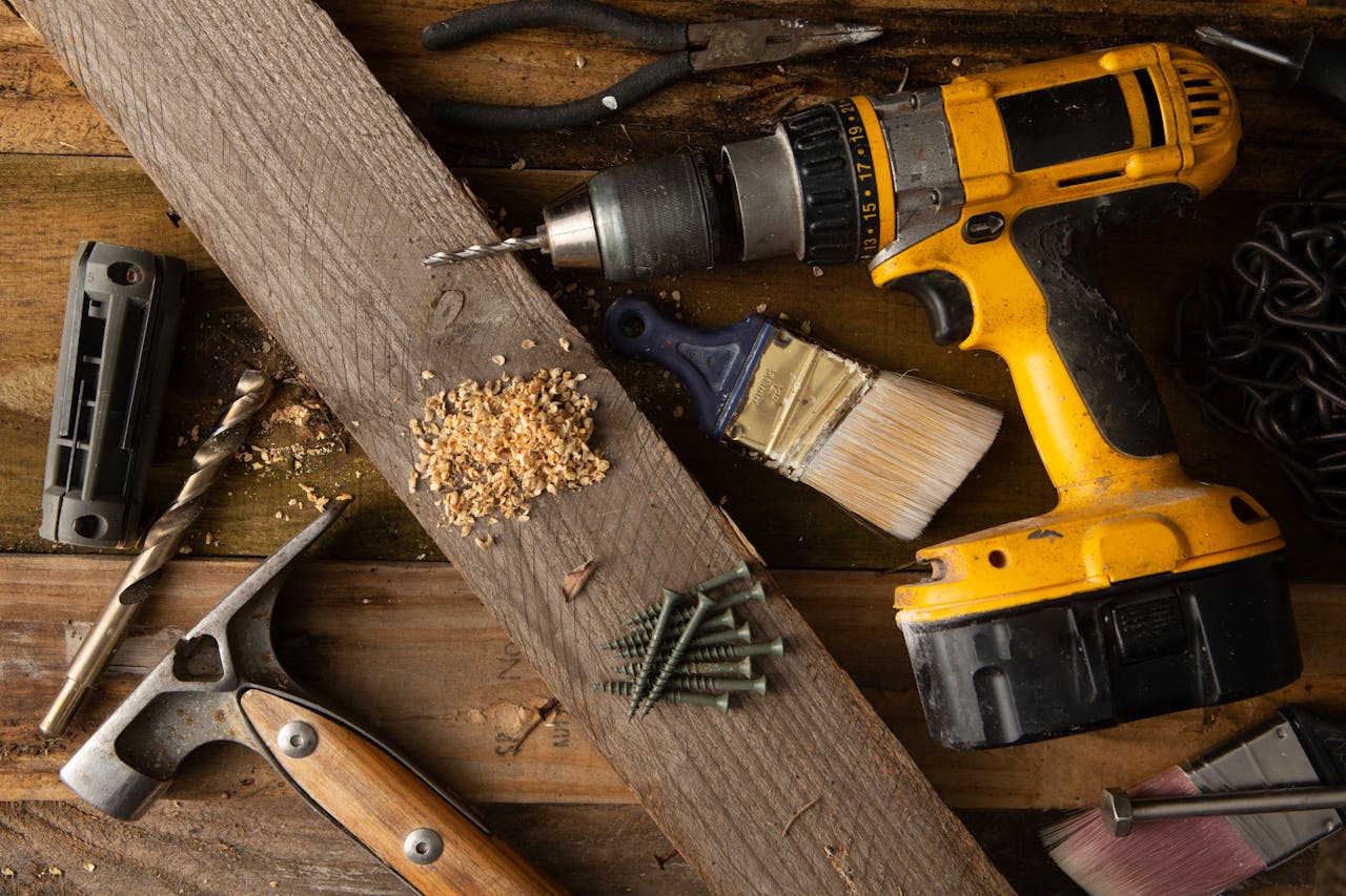 gallery-6 A close-up of various tools including a drill, hammer, and paintbrush on a wooden workbench.