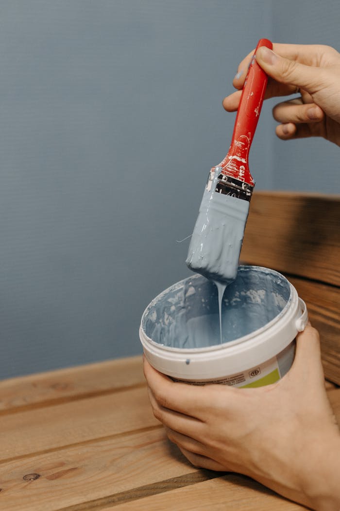 gallery-2 Hand holding a red-handled paintbrush dipped in sky blue paint over a wooden surface.